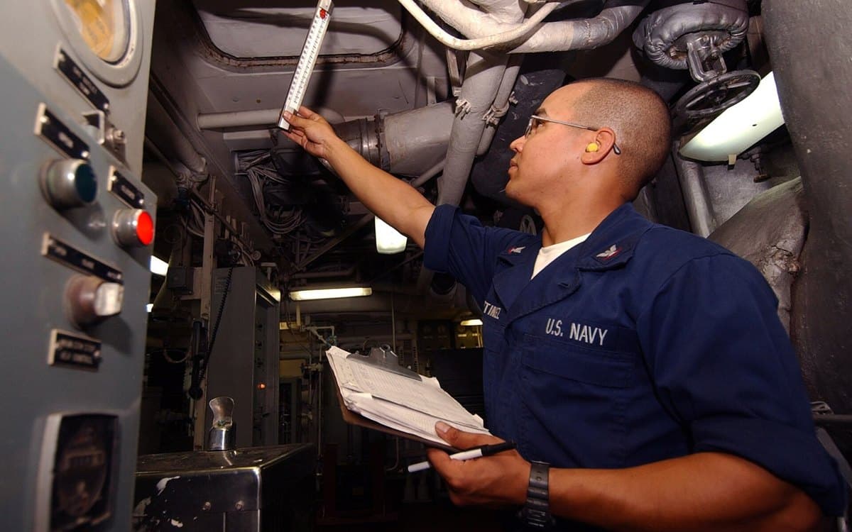A U.S. Navy sailor checking a gauge and logging readings in an engine room